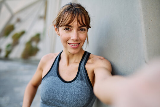 Young woman taking a selfie while posing against a wall after workout in athletic wear at urban location