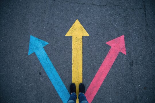 Top-down view of a person standing on asphalt with three painted arrows in blue, yellow, and red pointing in different directions