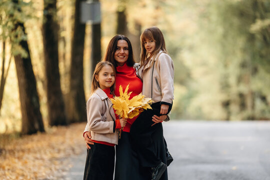 Smiling mother and two daughters holding autumn leaves together on a tree lined road in warm seasonal light, family portrait outdoors