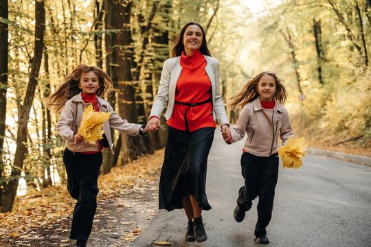 Mother walking with two daughters along a tree lined road while holding hands and autumn leaves in a sunlit forest setting