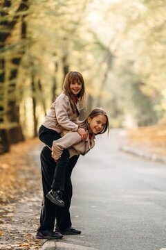 Two smiling girls sharing a playful piggyback ride on a quiet paved road surrounded by sunlit autumn trees in a wooded outdoor setting