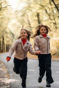 Two young girls holding hands while running along a tree lined road in autumn wearing matching beige jackets and red scarves outdoors