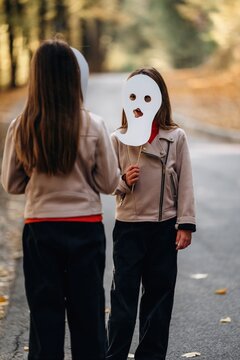 Two women in leather jackets facing each other on a tree lined road, one holding a blank white mask over her face in an outdoor portrait