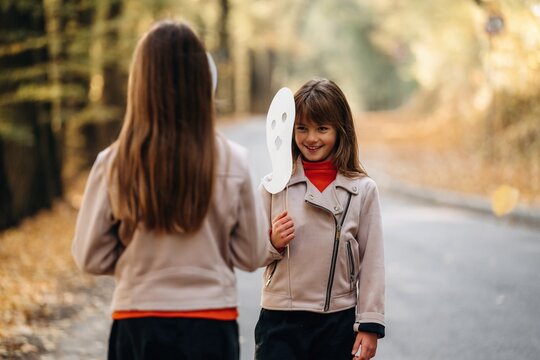 Two children outdoors with one holding a white mask while another faces her on a tree lined path during autumn, candid childhood scene