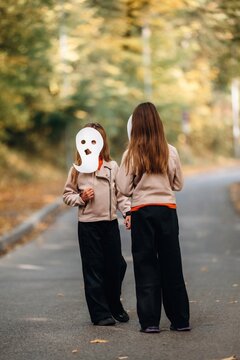 Two children in beige jackets walking on an autumn road, one wearing a white theatrical mask, viewed from behind in a wooded outdoor setting