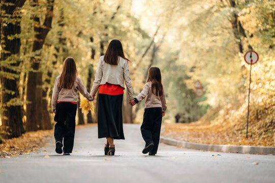 Woman walking hand in hand with two children along a tree lined road in autumn, viewed from behind in warm golden light and fallen leaves