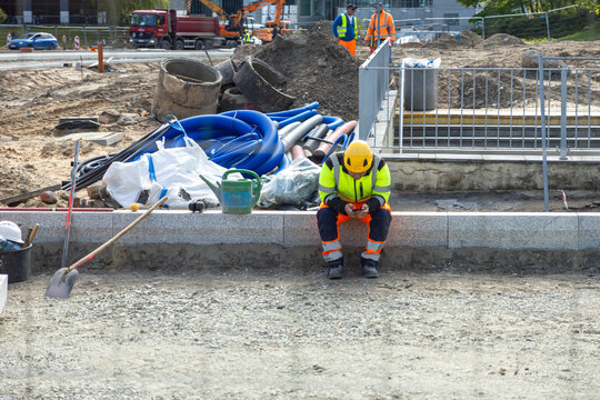 A construction worker sits on a concrete ledge using a smartphone during a break at an industrial site with pipes and heavy equipment visible in the background for civil engineering
