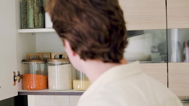 Man choosing food at open kitchen pantry with rice, lentils and couscous in glass jars. Food selection, pantry organization, grains, cereals and cooking ingredients. High quality 4k footage