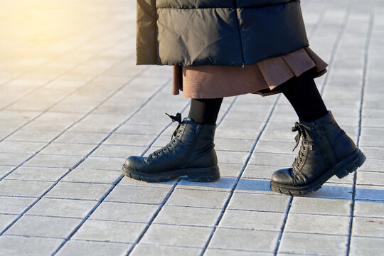 Woman in black leather boots strolling on paved tiles dusted with deicing salt, female legs in warm winter coat and black boots walking on street pavement covered in salt. Footwear care concept