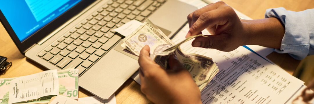 Header of African American adult counting cash money next to laptop, receipts and financial paperwork. Commercial concept representing personal budgeting, bookkeeping, banking and accounting