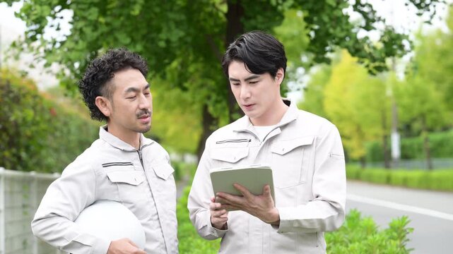 緑のある屋外でタブレットを確認しながら設備の不具合や点検箇所の問題に悩む2人の男性作業員、保守点検や現場トラブルのイメージ動画