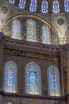 A wide calligraphic frieze stretches above illuminated arched stained glass windows and circular calligraphy medallions inside the Sultanahmet Blue Mosque in Istanbul, Turkey.