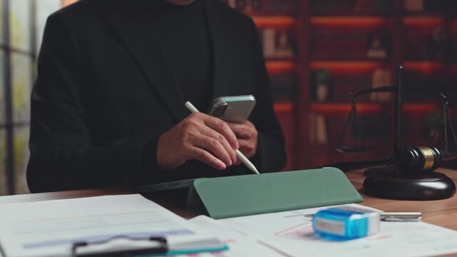 Lawyer writes on digital tablet holding smartphone near gavel balance scale in law firm office. Online legal consultation services, court justice contract review, corporate compliance management.