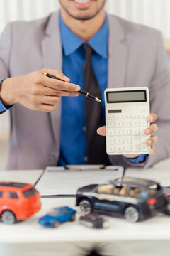 Smiling male auto dealer consultant sitting at desk presenting car models for vehicle sales contract.