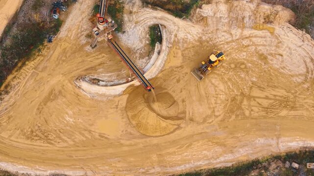 Aerial top down shot of sand quarry equipment. Conveyor belt drops sand onto a heap beside a yellow front loader on sandy ground.