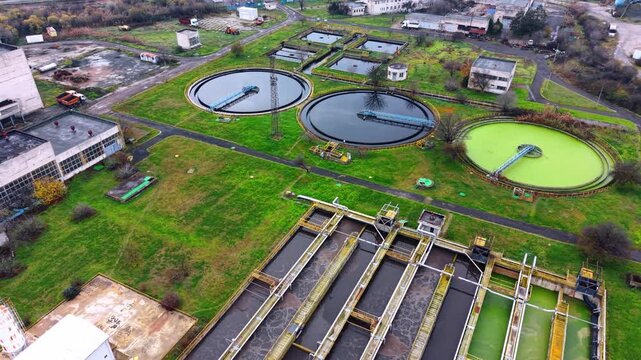 Aerial view of circular settling tanks and aeration basins. A drone perspective shows two large dark primary clarifiers and one green basin on grassy terrain.