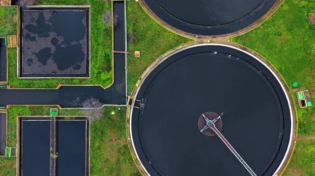Top down aerial shot of industrial water purification facility basins. Drone perspective captures rectangular channels and circular clarifier with dark liquid and green grass.