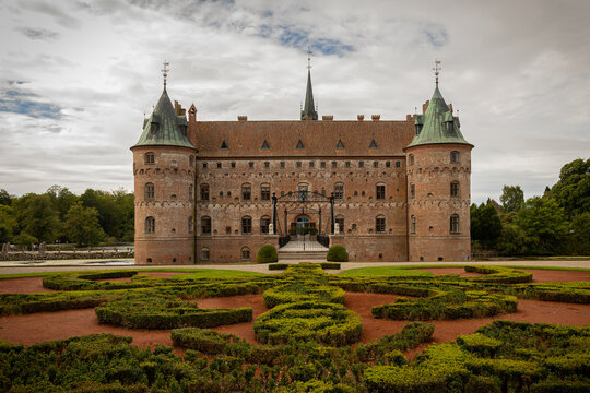A historic brick castle with two towers stands behind a symmetric formal garden featuring meticulously trimmed boxwood hedges under a soft, overcast cloudy sky.