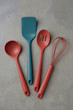 Flat lay of a teal silicone spatula and orange whisk, slotted spoon, and solid spoon on a textured grey surface.