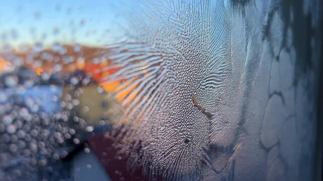Feathery frost crystals spread across window glass toward left side, revealing intricate branching patterns against blurred buildings, warm sunlight and clear blue sky