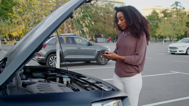 Concerned African American woman examines dead engine of car