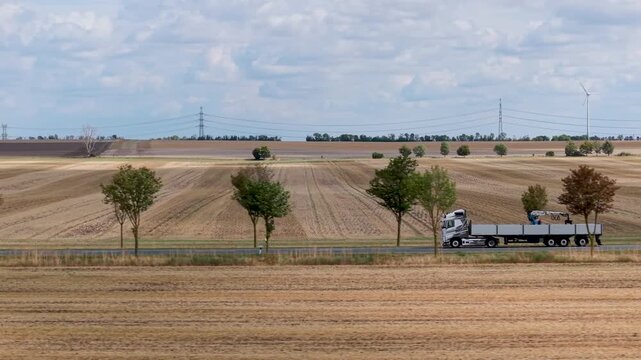 Aerial view follows flatbed truck with mounted crane along rural road beside harvested fields in Germany. Agricultural landscape stretches toward power lines and wind turbine beneath cloudy sky