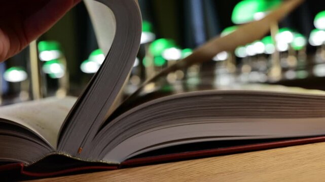 Hand turning pages of open book in library with rows of green banker lamps behind