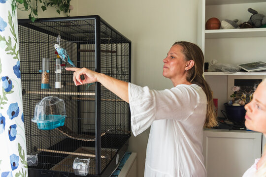 Mother Pointing at Pet Budgie in Cage with Daughter Watching