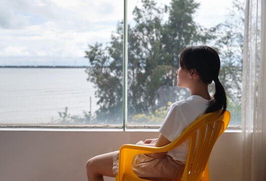 Chinese girl sits in front of a large window, looking out at the sea
