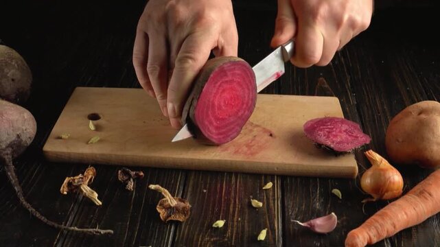 Hands slicing a red beet with a kitchen knife on a wooden cutting board surrounded by onions, carrots, potatoes, garlic, and vegetable scraps in a rustic kitchen setting