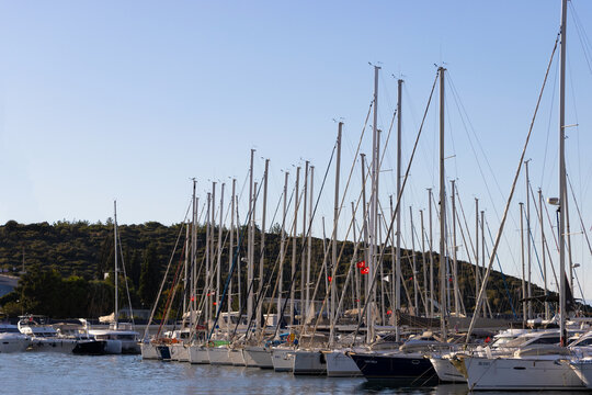 Scenic view of sailboats with tall masts docked at Teos Marina, their silhouettes contrasting against a clear blue sky and a lush green hillside. Sığacık, İzmir, Turkey. September 14, 2026.