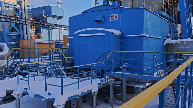Blue process unit 124 sits on elevated pipeline platform amid snow and steel railings. Insulated pipes and access walkways frame industrial plant scene, while quiet winter setting feels still.