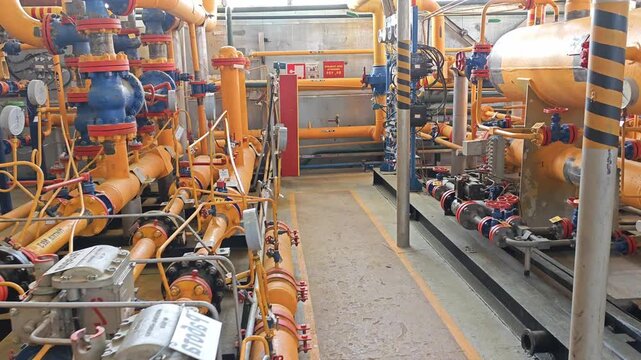 Orange valve system fills control room with dense pipework and gauges. Industrial equipment lines narrow aisles while blue fittings and red handwheels suggest active monitoring in busy plant interior.