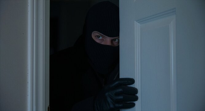 A masked burglar wearing a black balaclava and gloves peers cautiously through a slightly open door inside a dark room, conceptualizing home invasion, burglary, theft, and property crime.