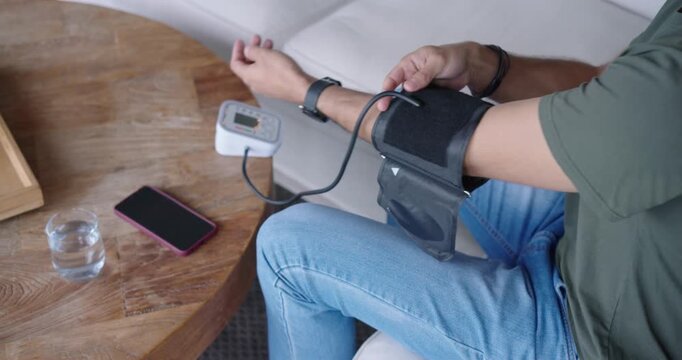 After pressing start, man is fastening cuff and operating monitor to measure blood pressure on sofa
