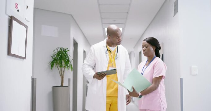 African American doctor and nurse reviewing patient chart and tablet in hospital corridor