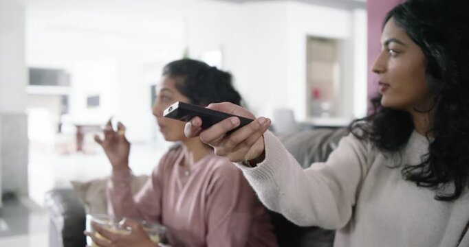 Indian mother and daughter couch watching TV, daughter using remote selecting, mother holding bowl
