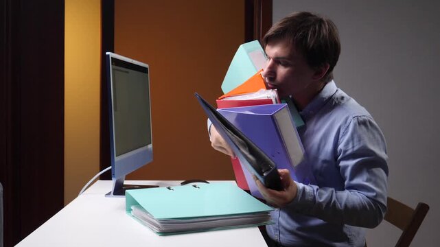 Office worker clutching colorful binders beside a computer, surrounded by stacked files and paperwork, conveying workplace stress, overload, burnout, and the pressure of multitasking