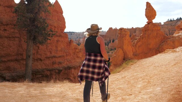 Back view female hiker trekking up dusty footpath among orange sandstone hoodoos with walking sticks, enjoying scenic journey through bryce canyon national park in utah state