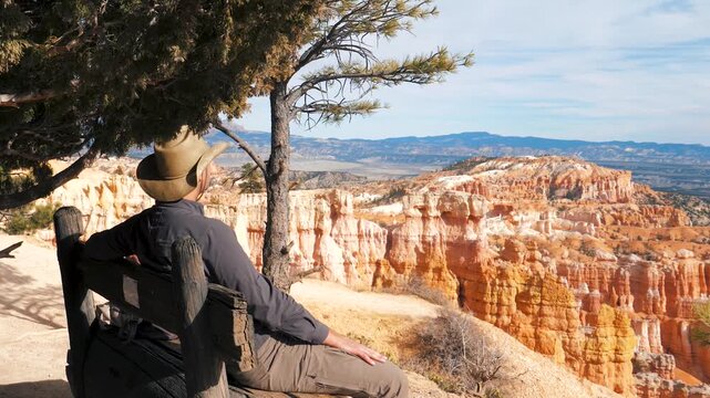 Male tourist wearing cowboy hat sitting on rustic wooden bench under trees, admiring colorful sandstone hoodoos and natural orange rock formations in bryce canyon national park in utah state