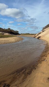 Shallow tidal inlet stream carving through golden sand dunes and coastal salt marsh under a sunny blue sky on Cape Cod, Massachusetts in vertical 4K