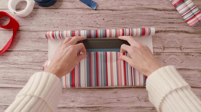 Woman's hands wrapping a book with a white, red and blue striped wrapping paper, and white, red and blue ribbons on wooden table. Real time video.