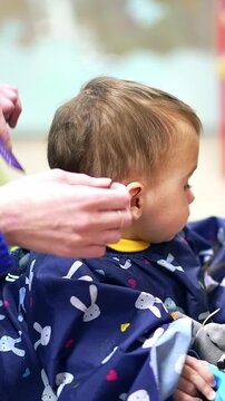 Barber combing toddler boy's hair picking the strand. Beautiful Caucasian baby having haircut at barber's. Blurred backdrop. Vertical video