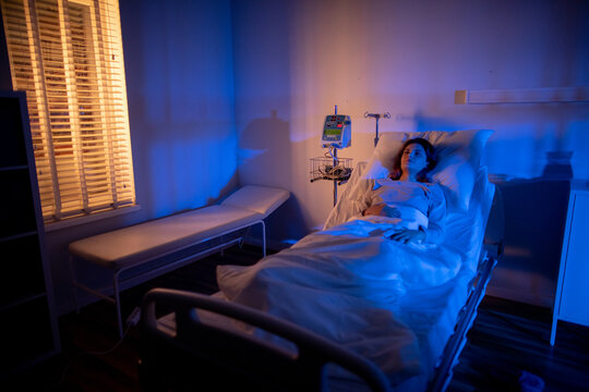 Hospital bed with infusion pump patient awake looking at mobile device, soft window light casting blinds pattern, empty guest chair and clinical equipment visible