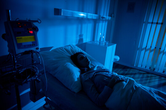 Patient lying under overnight observation in ward, distant monitor lights, IV pole silhouette, hush of nighttime, pale blue atmosphere, caregiver absence implied