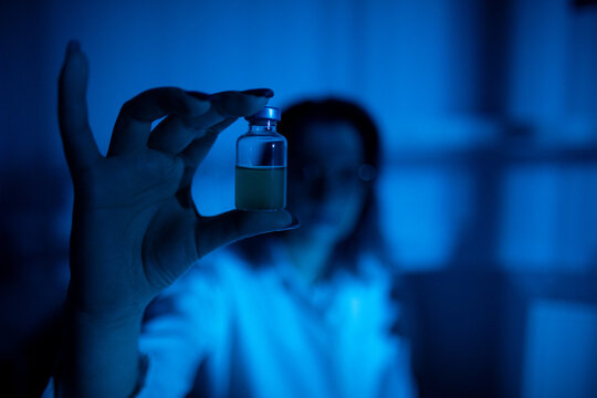 Laboratory environment with focus on sample inspection process. An intense scene capturing laboratory technician inspecting small vial carefully under blue lighting conditions