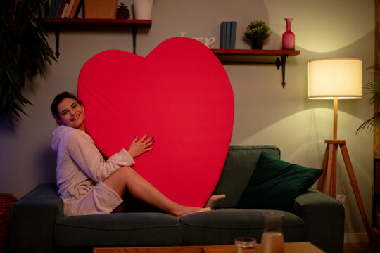 Joyful woman displaying love. Energetic woman offers oversized heart while relaxing at home. Happy woman presents giant red heart on modern furniture during cozy evening at home