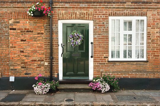 Traditional English cottage facade in Aylesbury, Buckinghamshire, featuring a dark green front door adorned with a floral wreath, exposed red brickwork, white sash windows, and colorful flower planter