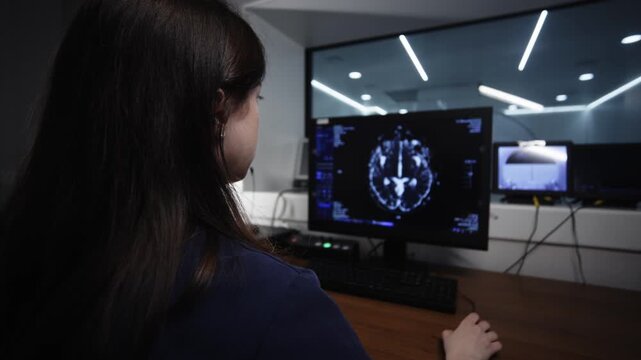 Radiologist viewing changing MRI brain scan images on computer monitor in dark diagnostic workstation through observation window in hospital imaging room