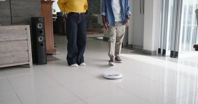 Diverse couple entering open living room, man crouching, touching white robot vacuum cleaning tiles